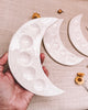 Hand holding a white ceramic moon-shaped plate with another plate in the background on a textured surface.