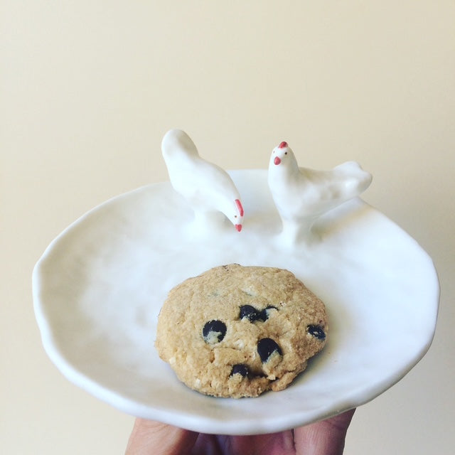 White ceramic dish with a cookie and two small chicken figurines on a beige background