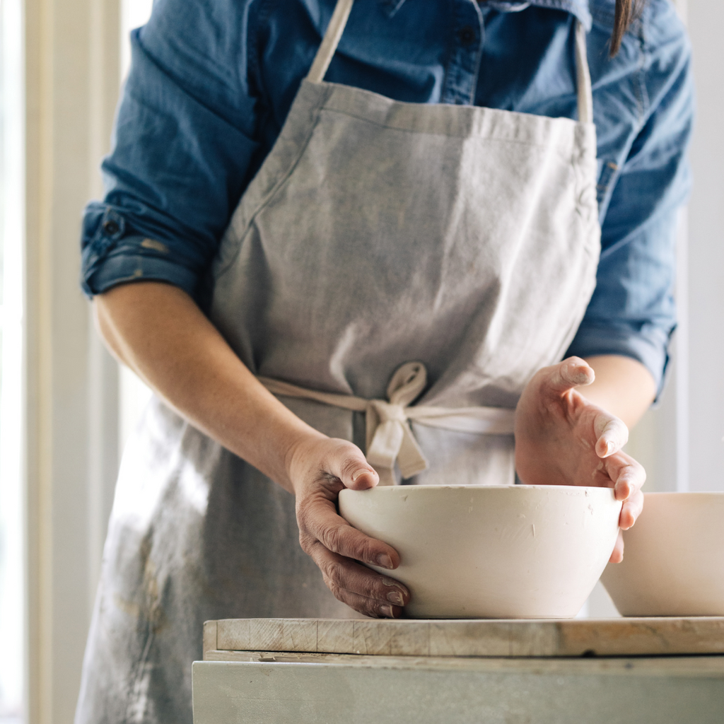 A person wearing a blue apron and an artist's hat is shown kneeling and working with clay in a wheel throwing technique.