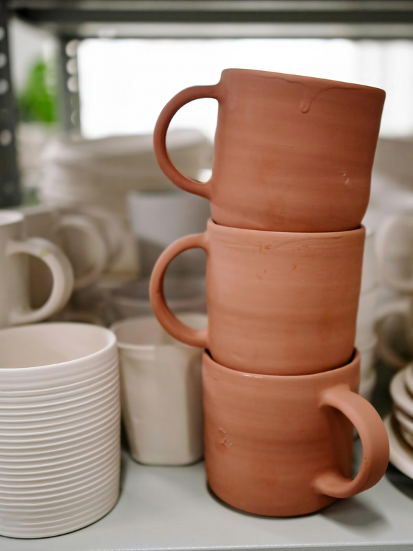 Stack of terracotta mugs on a surface with blurred ceramic items in the background