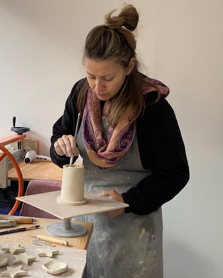 A woman in a pottery studio wearing a black long-sleeve shirt, a gray apron, and a purple patterned scarf, sculpting a small clay vessel on a pottery wheel.