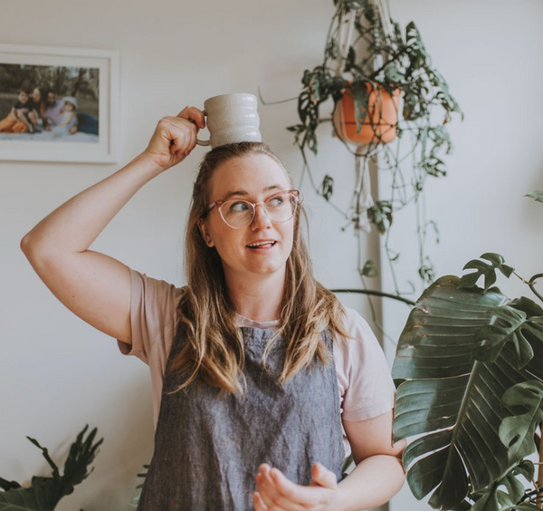 Woman holding a mug above her head in a home setting with plants and family photos.
